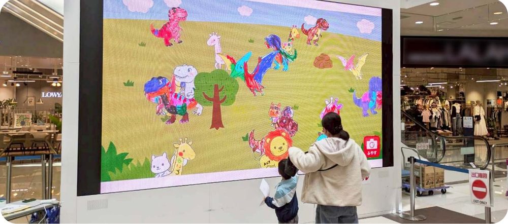 大型モニターに映った恐竜のぬりえを見る親子