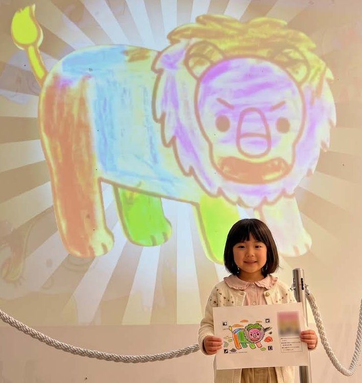 大画面に映ったライオンのぬりえの前で記念撮影する女の子